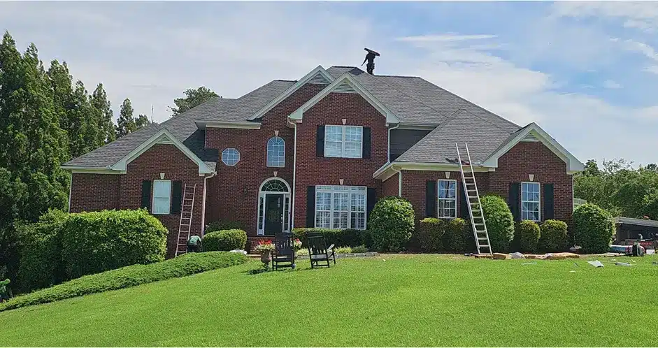 Roofer standing on top of large brick house during active roof repair process with ladders positioned on both sides by Skyline Contracting