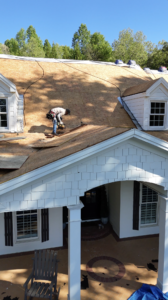 Certified roofing contractors Gainesville, GA working on roof renovation with exposed wooden sheathing, dormer windows, and white siding, using tools to secure new materials during repair.