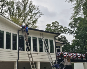 A technician standing on a tall extension ladder installing a dark seamless gutter system on a white multi-windowed house to address common new gutters signs gutter installation service in Gainesville by Skyline Contracting