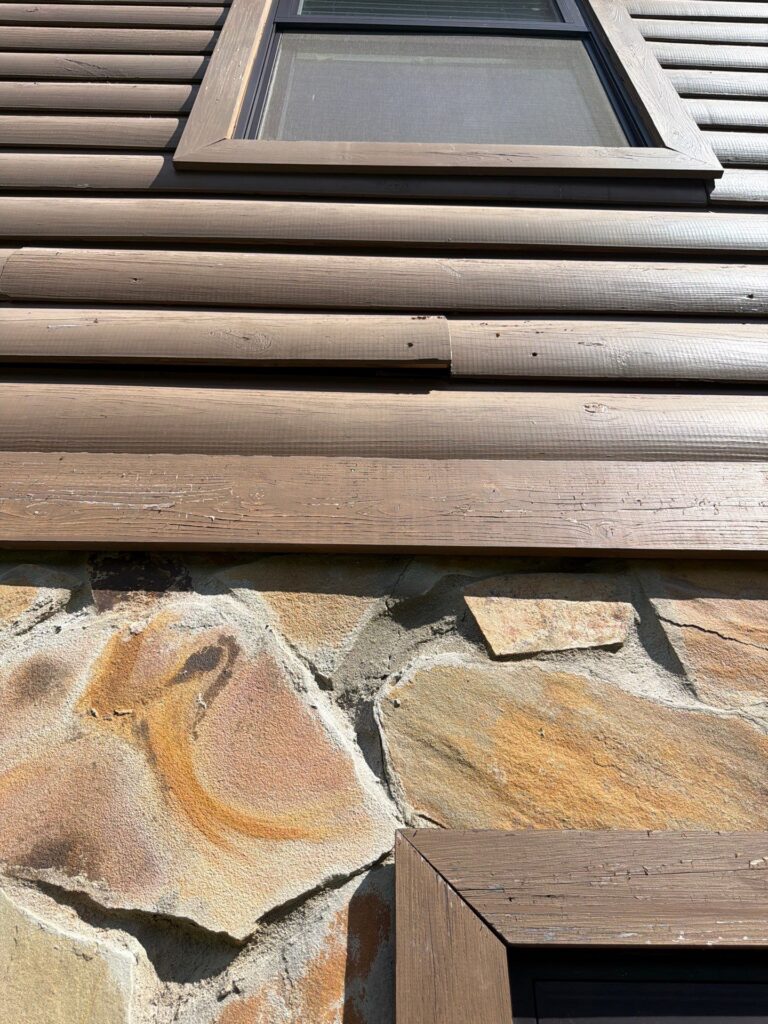 Aged brown wood-look siding and stone veneer exterior wall showing gaps near a window during a house siding replacement process siding replacement service in Gainesville by Skyline Contracting